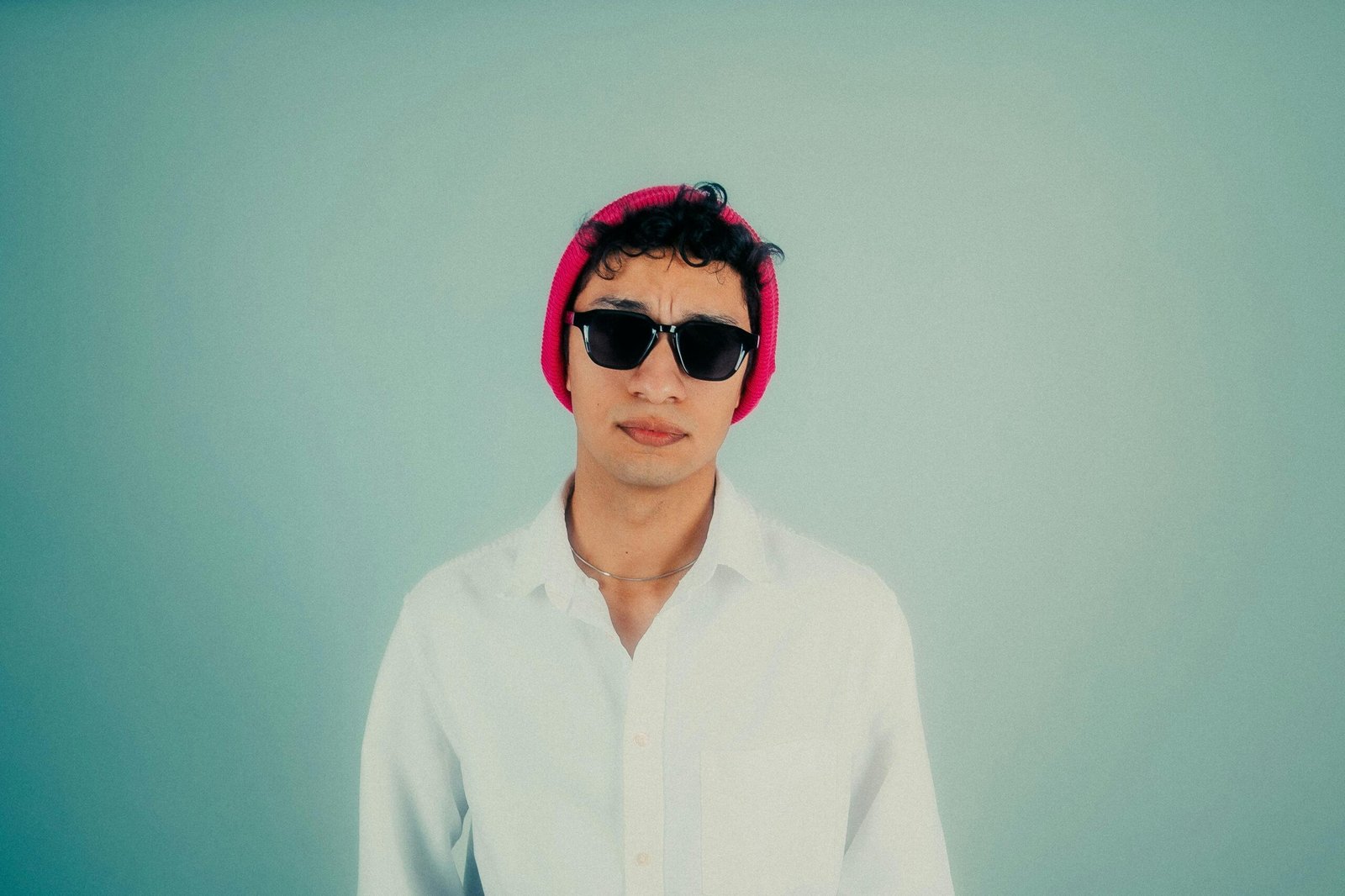 Fashionable young man wearing sunglasses and a pink beanie, set against a minimalist pastel background.